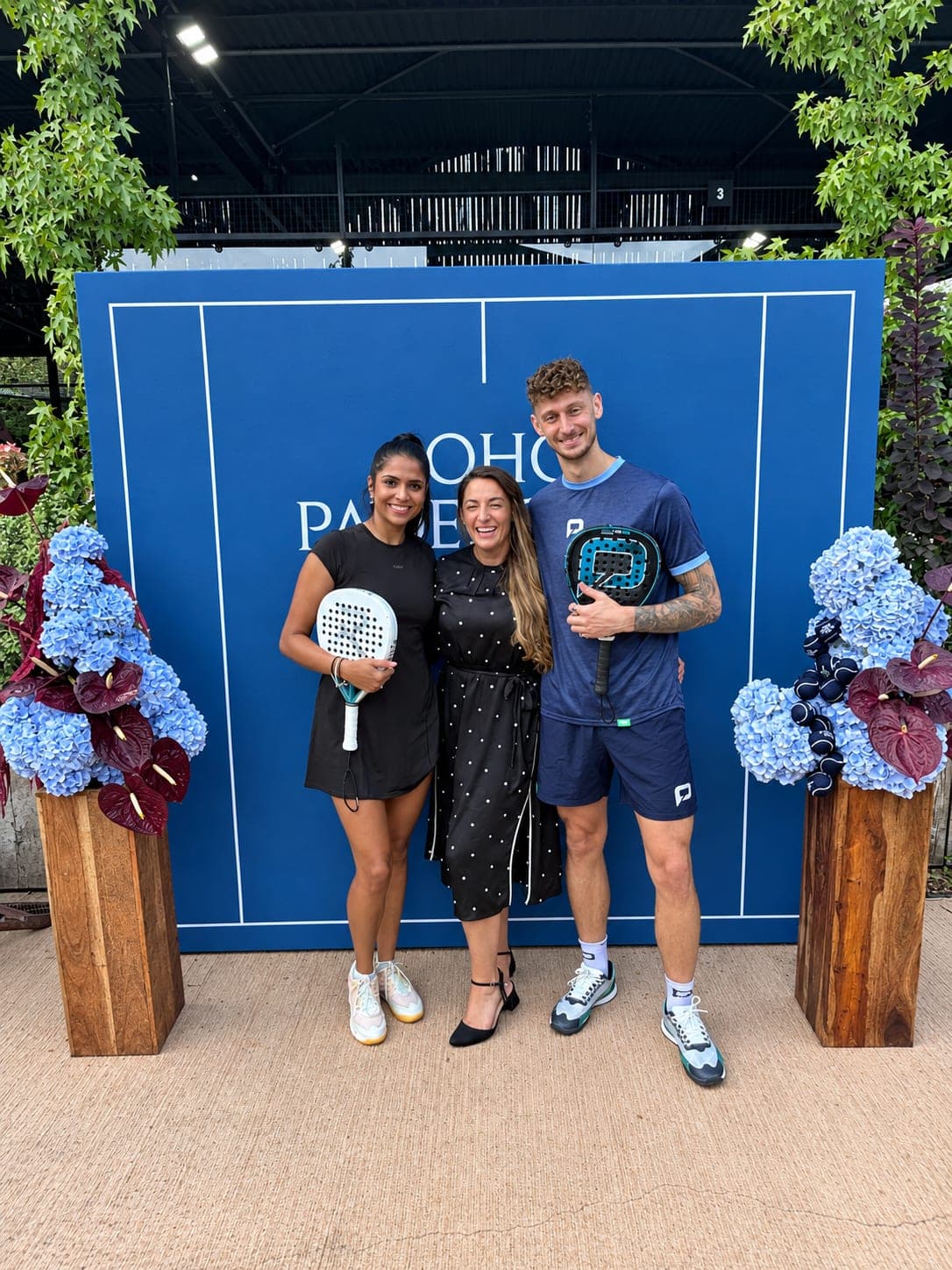 Two padel players holding their rackets, with a colleague between them, smiling in front of a blue court-lines backdrop at a padel event.